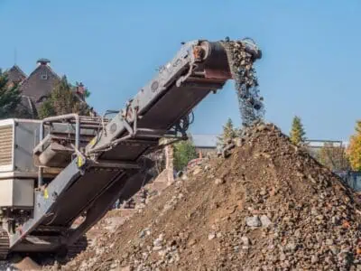 Excavator with rock conveyor belt unloading soil and rubble onto a large pile at a construction site